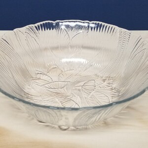 May include: A clear glass serving bowl with a scalloped edge and a floral leaf design. The bowl is round with a wide opening. It is set on a light-coloured surface against a dark blue backdrop.