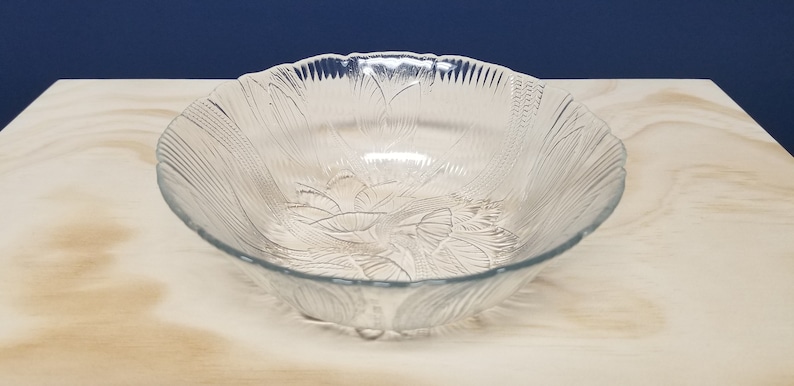 May include: A clear glass serving bowl with a scalloped edge and a floral leaf design. The bowl is round with a wide opening. It is set on a light-coloured surface against a dark blue backdrop.