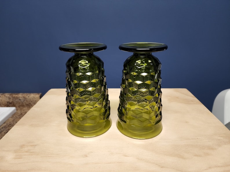 May include: Two olive green glass goblets standing upside down on their rims. Each goblet features a distinct geometric honeycomb pattern across the body, a smooth circular base at the top, and a smooth circular rim at the bottom.