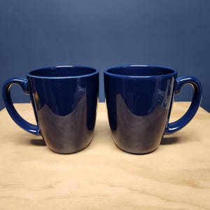 May include: Two blue ceramic mugs with handles. The mugs are on a wooden surface.