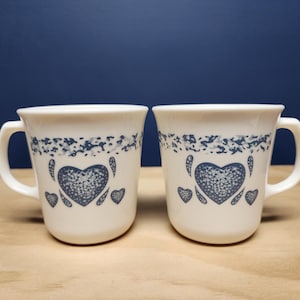 May include: Two white ceramic mugs with blue heart and floral designs. Each mug has a handle and a blue floral pattern near the rim. The mugs are on a wooden surface with a dark blue background.