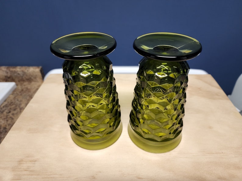 May include: Two matching olive green glass tumblers featuring a textured, geometric diamond pattern across the exterior. Each glass has a wide, smooth circular base and a slightly flared, smooth rim, showcasing a translucent finish throughout.