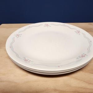 May include: A stack of white dinner plates featuring a delicate pattern along the rim. The design consists of thin, wavy light blue lines accented by small clusters of pink and green floral motifs spaced evenly around the circular edge of the plates.