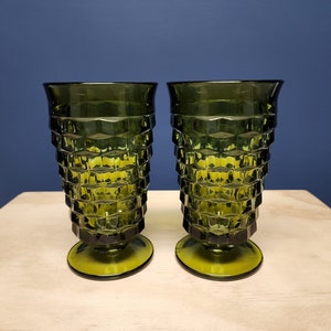 May include: Two matching olive green glass tumblers featuring a textured, geometric honeycomb pattern on the exterior. Each glass has a flared rim, a tapered body, and a circular pedestal base, creating a distinct silhouette for these drinking vessels.