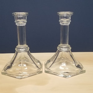 May include: Two clear glass candle holders. Each has a square base that tapers to a long, slender stem, and a small, round top to hold a candle. The candle holders are displayed on a light-colored wooden surface against a dark blue background.