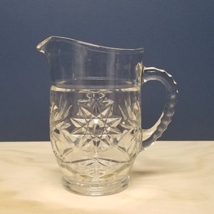 May include: A clear glass pitcher with a star-shaped pattern, filled with water, on a light wood surface.