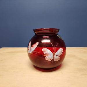 May include: A round, deep red glass vase featuring hand-painted white butterflies with delicate wing details. The vase has a wide, circular opening at the top and a smooth, translucent finish that allows light to pass through the colored glass surface.