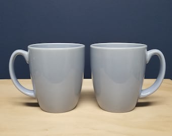 May include: Two gray ceramic mugs with handles on a light wood surface.