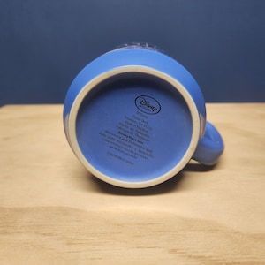 Puede incluir: Una taza de cer&aacute;mica azul con interior blanco, vista desde abajo. La taza tiene un asa y una base circular. La parte inferior de la taza presenta texto y el logotipo de Disney Store.