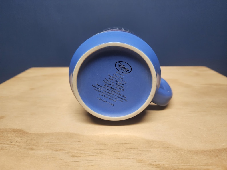 Puede incluir: Una taza de cer&aacute;mica azul con interior blanco, vista desde abajo. La taza tiene un asa y una base circular. La parte inferior de la taza presenta texto y el logotipo de Disney Store.