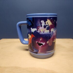 Puede incluir: Una taza de cer&aacute;mica con asa y borde azules, con un dise&ntilde;o de dibujos animados colorido. El dise&ntilde;o incluye varios personajes en un entorno caprichoso. La taza est&aacute; colocada sobre una superficie de madera sobre un fondo azul oscuro.