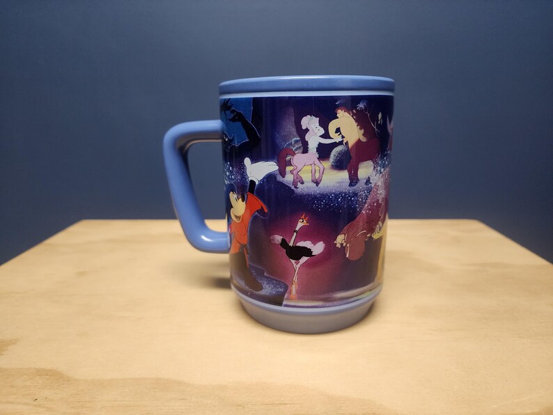 Puede incluir: Una taza de cer&aacute;mica con asa y borde azules, con un dise&ntilde;o de dibujos animados colorido. El dise&ntilde;o incluye varios personajes en un entorno caprichoso. La taza est&aacute; colocada sobre una superficie de madera sobre un fondo azul oscuro.