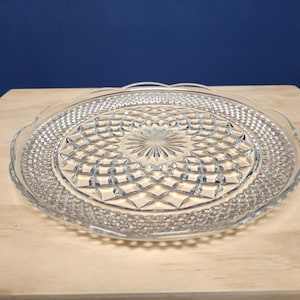May include: A clear, round glass cake plate with a decorative diamond pattern and scalloped edges. The plate is approximately 12 inches in diameter. The plate is on a light-colored wooden surface with a dark blue background.