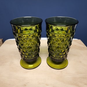 May include: Two matching olive green glass tumblers featuring a faceted, geometric diamond pattern on the exterior. Each glass has a wide, flared rim that tapers down to a circular pedestal base, creating a distinct, textured silhouette.