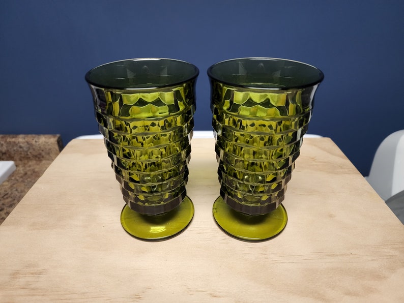 May include: Two matching olive green glass tumblers featuring a faceted, geometric diamond pattern on the exterior. Each glass has a wide, flared rim that tapers down to a circular pedestal base, creating a distinct, textured silhouette.