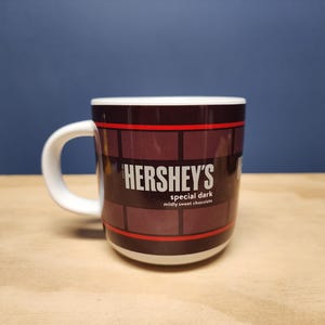 May include: A white-handled mug with a dark brown brick pattern and red stripes. The mug features the Hershey's logo and the text "special dark mildly sweet chocolate". The mug is on a wooden surface.