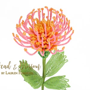 May include: A beaded flower with pink and orange petals and green leaves. The flower is made of small beads and is a realistic representation of a real flower. The flower is on a white background. Bead & Blossom by Lauren Harpster.
