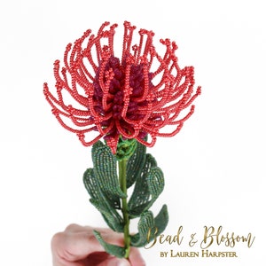 May include: A beaded red and green protea flower, a type of South African flower, with the text "Bead & Blossom by Lauren Harpster" at the bottom of the image.