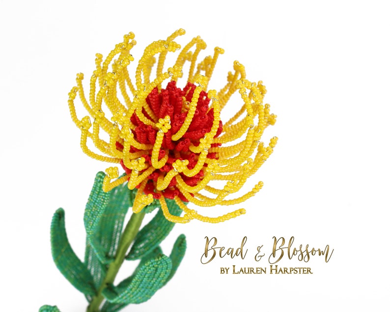 May include: A beaded flower sculpture in the shape of a red and yellow protea. The flower is made with small beads and wire. The sculpture is on a white background. Bead & Blossom by Lauren Harpster.