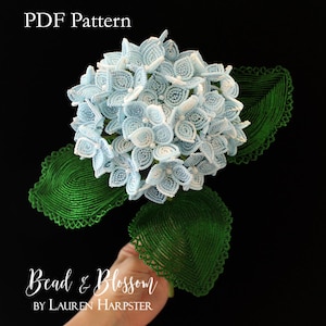 May include: A beaded hydrangea flower sculpture featuring light blue and white petals with three dark green serrated leaves. The text PDF Pattern Bead & Blossom by Lauren Harpster is visible on the image against a solid black background.