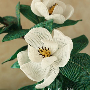 May include: A close-up of a white beaded magnolia flower with a yellow center. The flower is part of a larger beaded floral arrangement with green leaves. The text "Bead & Blossom by Lauren Harpster" is visible in the bottom right corner of the image.