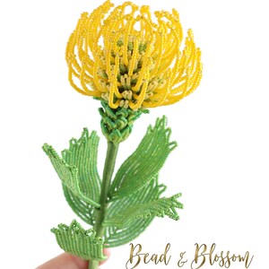 May include: A beaded flower with yellow petals and green leaves. The flower is held in a hand and the text "Bead & Blossom by Lauren Harpster" is visible below the flower.