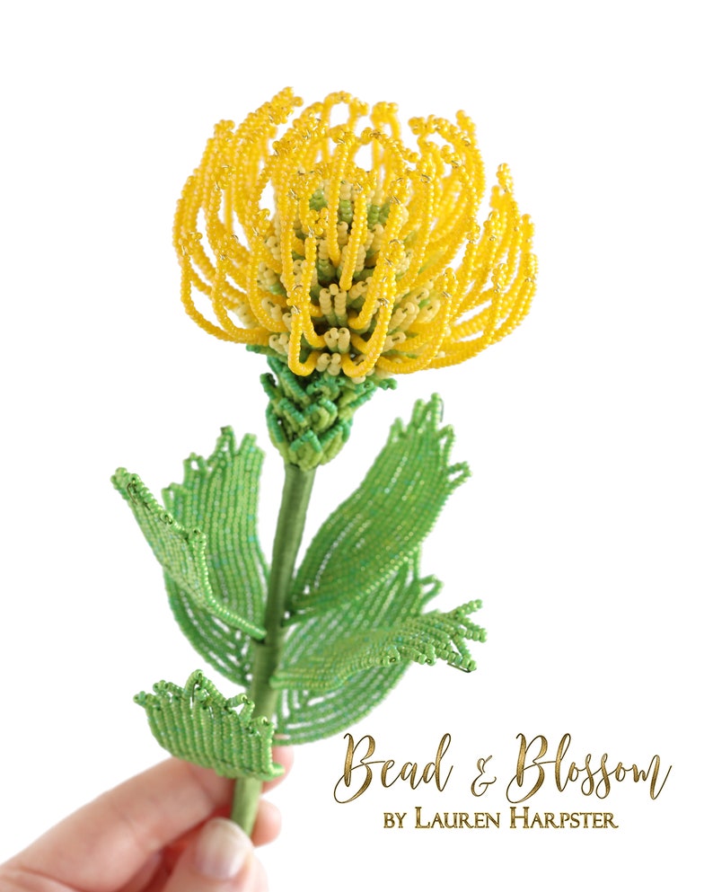 May include: A beaded flower with yellow petals and green leaves. The flower is held in a hand and the text "Bead & Blossom by Lauren Harpster" is visible below the flower.