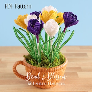 May include: A bouquet of beaded crocus flowers in purple, white, and yellow colors, arranged in a brown ceramic pot with green moss. The pot has a handle and the text "Bead & Blossom by Lauren Harpster" is printed on the front.