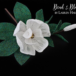 May include: A white beaded magnolia flower with green beaded leaves. The flower is in bloom and has a yellow center. The text "Bead & Blossom by Lauren Harpster" is in the upper right corner of the image.