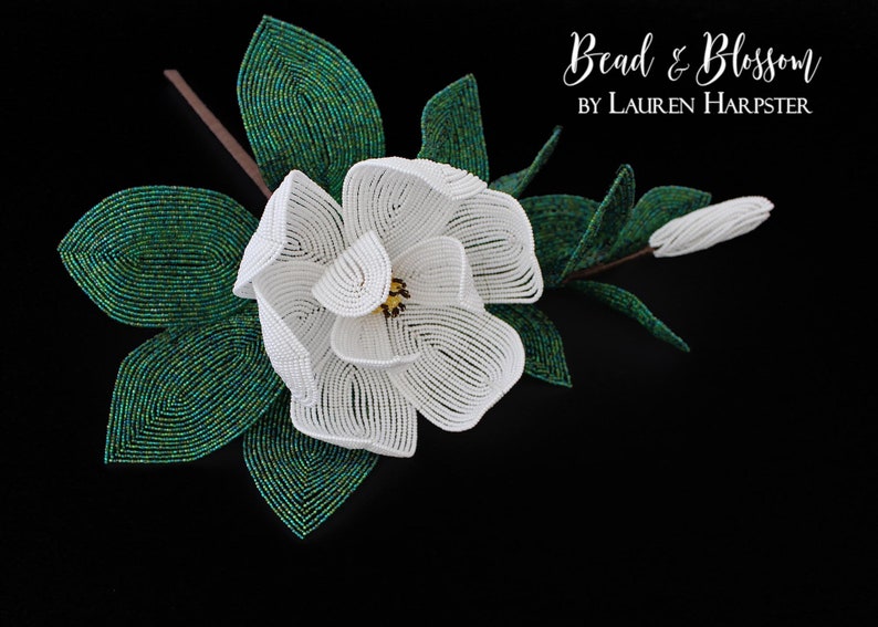 May include: A white beaded magnolia flower with green beaded leaves. The flower is in bloom and has a yellow center. The text "Bead & Blossom by Lauren Harpster" is in the upper right corner of the image.