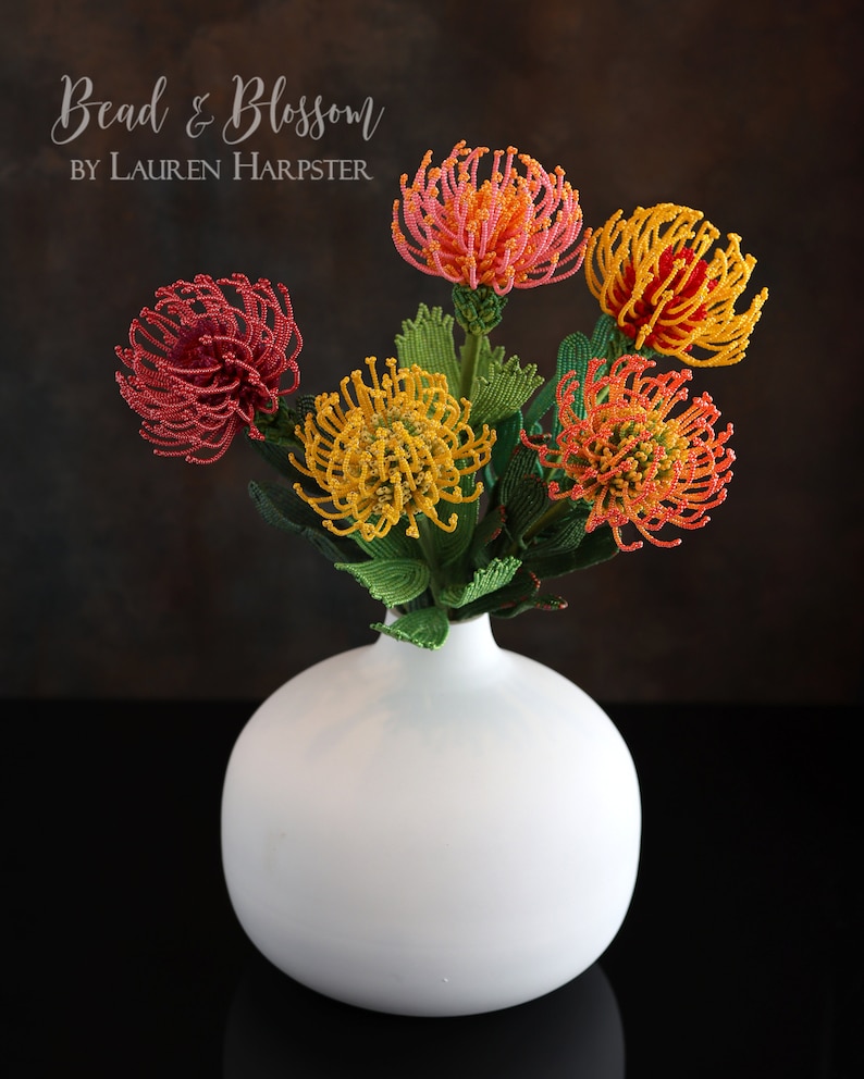 May include: A bouquet of beaded flowers in a white vase. The flowers are red, orange, and yellow and have a spiky, pinecone-like shape. The text "Bead & Blossom BY LAUREN HARPSTER" is visible at the top of the image.