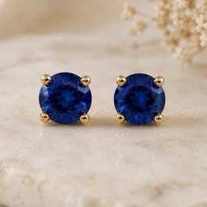 Puede incluir: Un par de pendientes de botón con zafiros azules de talla redonda montados en engastes de oro de cuatro garras. Cada pendiente presenta una gema facetada de color azul intenso y vibrante sujeta por cuatro pequeñas garras doradas.