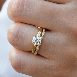 Peut inclure: Gros plan sur une main de femme portant deux bagues en or. La bague du haut présente un gros diamant rond et clair. La bague du bas est une bande délicate avec de petits diamants ronds.