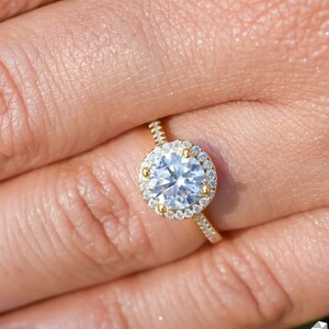 May include: A gold engagement ring with a large round center stone surrounded by a halo of smaller diamonds. The ring is on a finger with light purple nail polish.