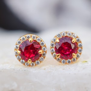 May include: A pair of gold earrings with a red gemstone surrounded by a halo of smaller white gemstones.