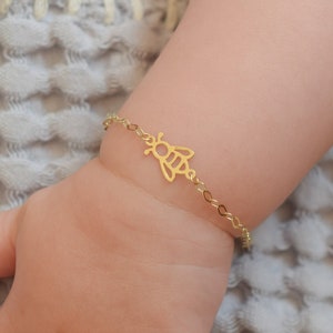 Könnte beinhalten: Ein goldenes Armband mit einem Bienen-Anhänger am Handgelenk eines Babys. Das Armband ist zart und hat ein schlichtes Design.