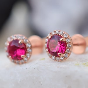 Op de afbeelding: Een paar oorbellen met een ronde robijn-edelsteen in een halo van kleinere witte edelstenen, gemaakt van roségoud.