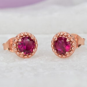 May include: A pair of rose gold stud earrings with a small, round, deep red gemstone in the center of each earring. The earrings have a beaded border around the gemstone.