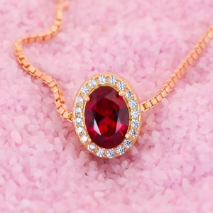 May include: A delicate gold chain necklace featuring a red oval gemstone set in a halo of smaller clear gemstones.