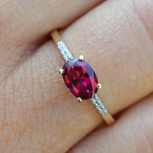 Könnte beinhalten: Ein goldener Ring mit einem roten ovalen Edelstein, der von kleineren weißen Edelsteinen umgeben ist.