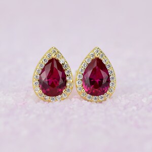Könnte beinhalten: Ein Paar goldene Ohrringe mit einem tropfenförmigen Rubin-Edelstein, der von einem Heiligenschein aus kleineren, klaren Edelsteinen umgeben ist.