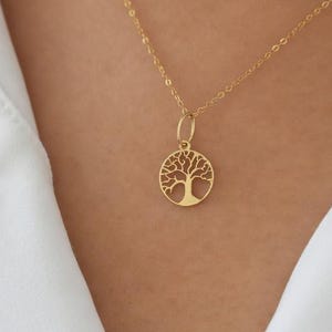 May include: A gold-colored necklace with a tree of life pendant. The pendant is circular with a detailed tree design. The necklace is worn over a white collared shirt.