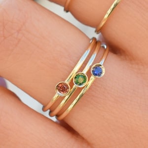 Peut inclure: Quatre bagues en or avec de petites pierres précieuses. Les bagues sont empilées sur un doigt. Les pierres précieuses sont bleues, vertes, orange et jaunes.