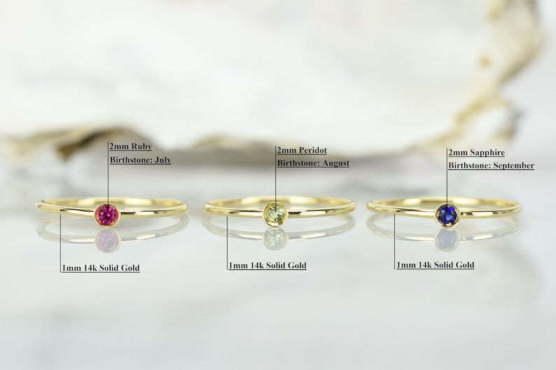 May include: Three gold rings with different colored gemstones. The rings are 1mm thick and made of 14k solid gold. The gemstones are 2mm in diameter and are ruby, peridot, and sapphire. The text below each ring reads "2mm Ruby Birthstone: July", "2mm Peridot Birthstone: August", and "2mm Sapphire Birthstone: September".