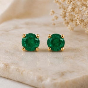 Puede incluir: Un par de pendientes de botón con piedras preciosas redondas de color verde vibrante en engastes de metal dorado de cuatro puntas. Cada pendiente presenta una piedra circular facetada asegurada por cuatro pequeñas puntas de oro pulido.