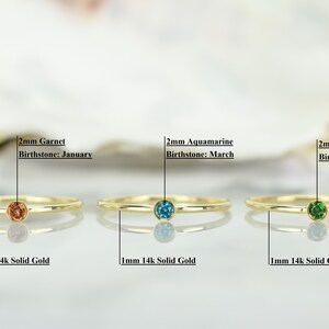 May include: Three gold rings with different colored gemstones. The rings are 1mm thick and made of 14k solid gold. The gemstones are 2mm in diameter and are garnet, aquamarine, and emerald. The text below each ring reads "1mm 14k Solid Gold" and the text above each ring reads "2mm Garnet Birthstone: January", "2mm Aquamarine Birthstone: March", and "2mm Emerald Birthstone: May".