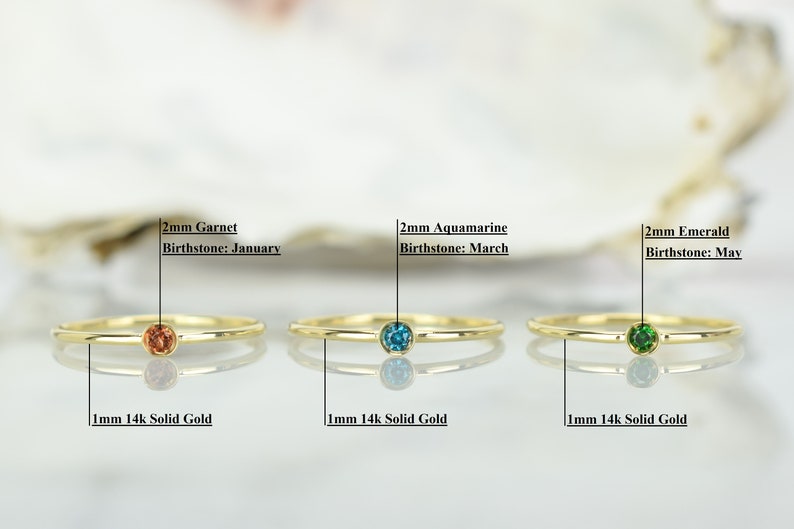 May include: Three gold rings with different colored gemstones. The rings are 1mm thick and made of 14k solid gold. The gemstones are 2mm in diameter and are garnet, aquamarine, and emerald. The text below each ring reads "1mm 14k Solid Gold" and the text above each ring reads "2mm Garnet Birthstone: January", "2mm Aquamarine Birthstone: March", and "2mm Emerald Birthstone: May".