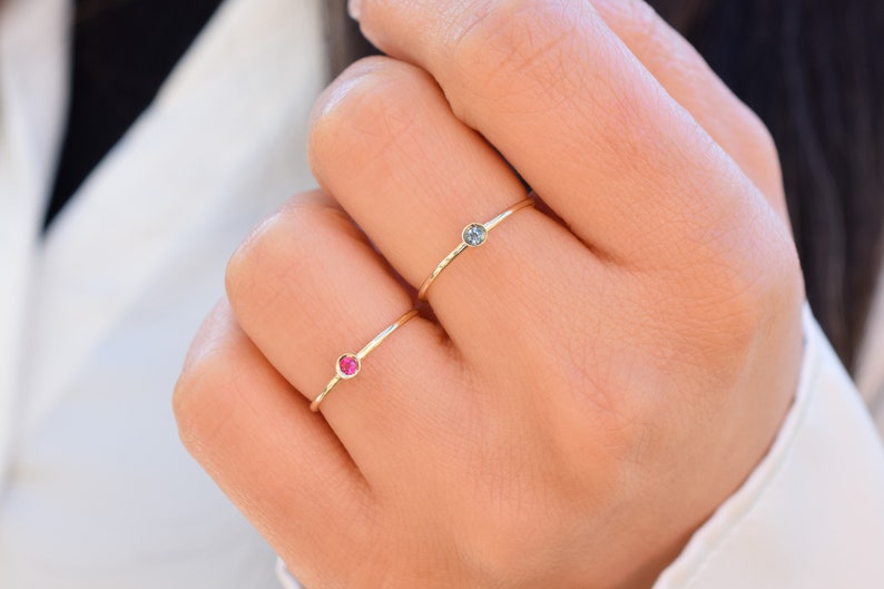 May include: Two delicate gold rings with small gemstones. One ring has a red gemstone and the other has a blue gemstone.