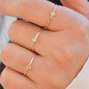 May include: Three gold rings with small gemstones on a person's finger. The rings are simple and delicate, with a single stone on each ring. The stones are different colors, one is clear, one is yellow, and one is a light brown.