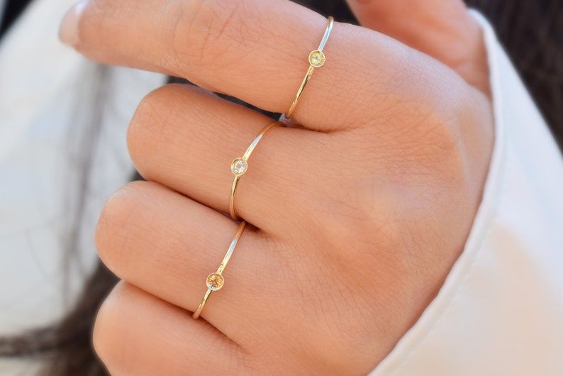 May include: Three gold rings with small gemstones on a person's finger. The rings are simple and delicate, with a single stone on each ring. The stones are different colors, one is clear, one is yellow, and one is a light brown.